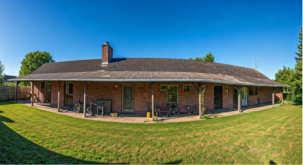 Long brick house with a covered porch and chimney. Bicycles and tools are visible. The lush green lawn and blue sky create a calm vibe.